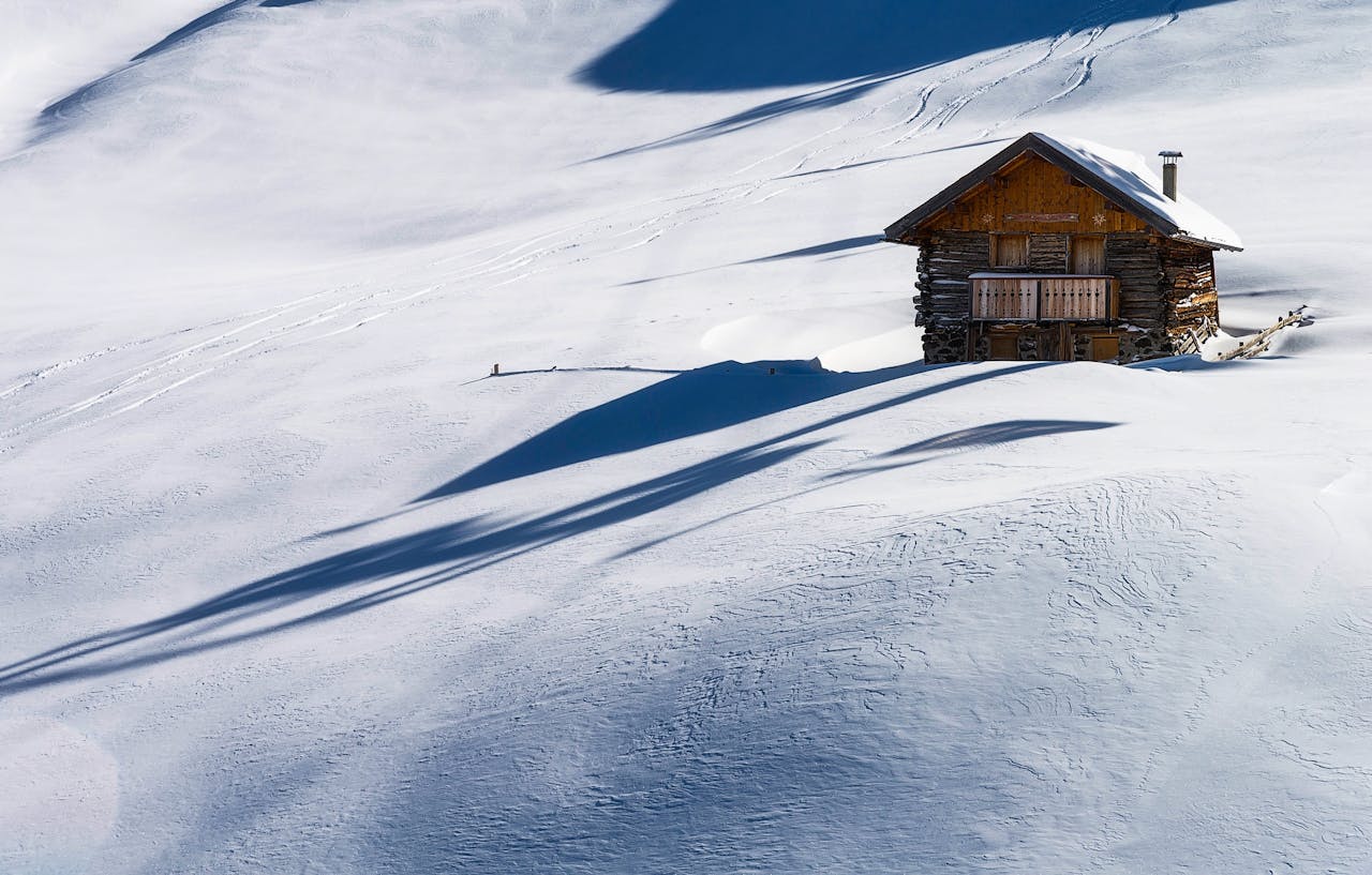 A secluded wooden cabin surrounded by a pristine snowy landscape under clear blue skies.