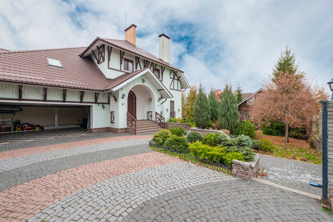 A quaint suburban house with a beautiful garden and brick driveway under a partly cloudy sky.